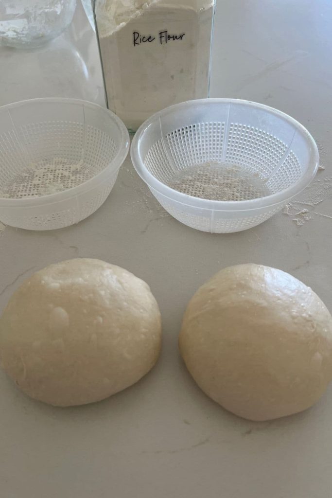 Two shaped sourdough bread bowls sitting in front of two plastic ricotta baskets. There is a glass jar of rice flour in the background.
