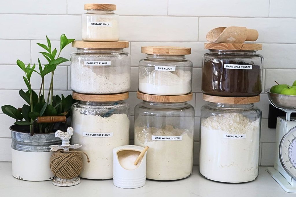 3 large jars of flour surrounded by some smaller jars containing rye flour, dark malt flour, diastatic malt and salt.