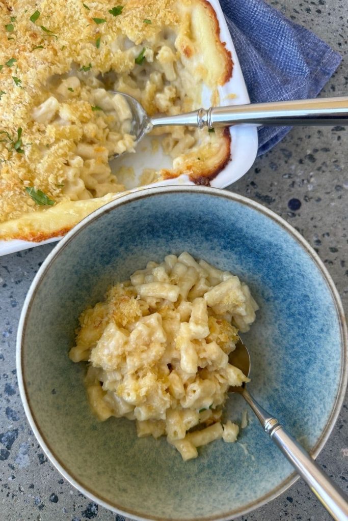 Sourdough mac and cheese served in a blue stoneware bowl. You can see the rest of the mac and cheese in a white baking dish behind the individual serving.