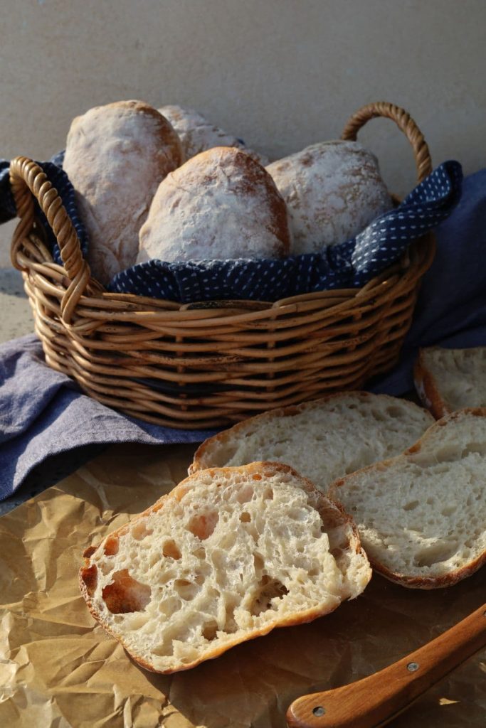 A basket of sourdough cibatta rolls. There is a cibatta roll that has been sliced in half sitting in front of the basket.