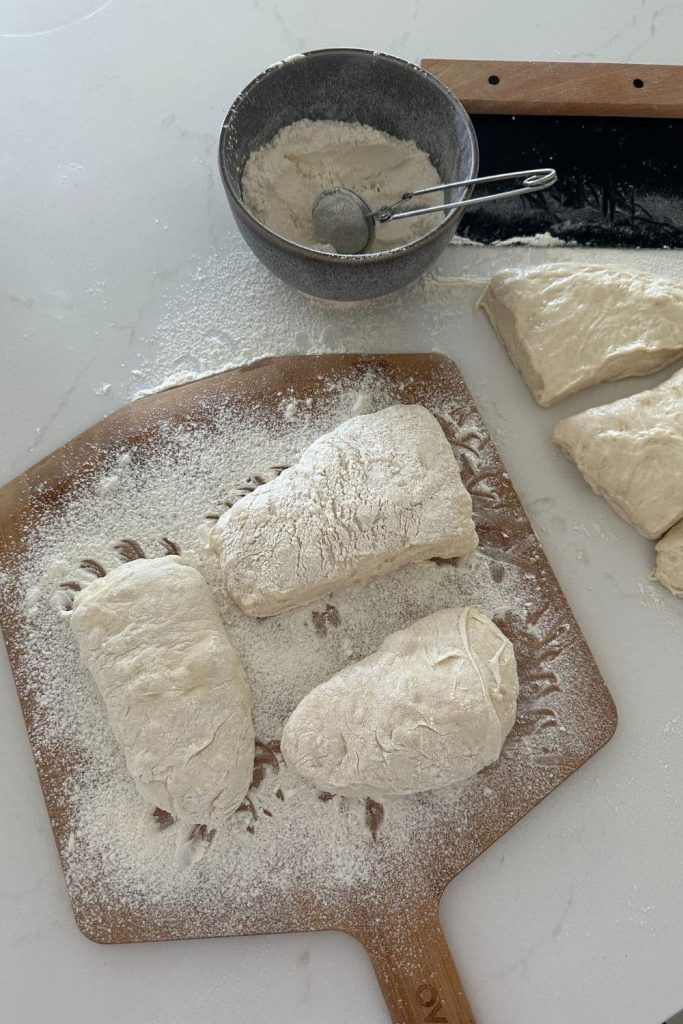sourdough cibatta dough sitting on a heavily floured pizza peel. You can see a bowl of flour with a tea strainer sitting in the photo, along with a black steel dough scraper.