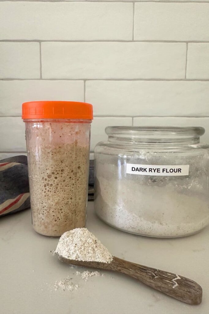 A jar of rye sourdough starter with an orange lid sitting next to a large glass canister of dark rye flour. There is a wooden spoon in front of these jars with some dark rye flour on it.