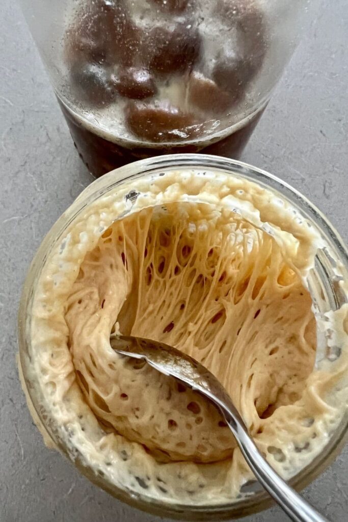 Looking down into the top of a jar of bubbly sourdough starter. There is a a metal spoon pulling the starter back from the side of the jar so you can see the bubbles underneath. You can see a jar of yeast water made from raisins at the top of the photo.
