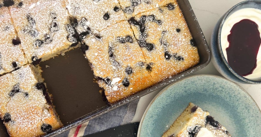 A photo of a tray of sourdough sheet pan pancakes with a piece missing from the tray. The piece of pancake is sitting on a blue plate to the right of the tray, garnished with vanilla yogurt and berry coulis.