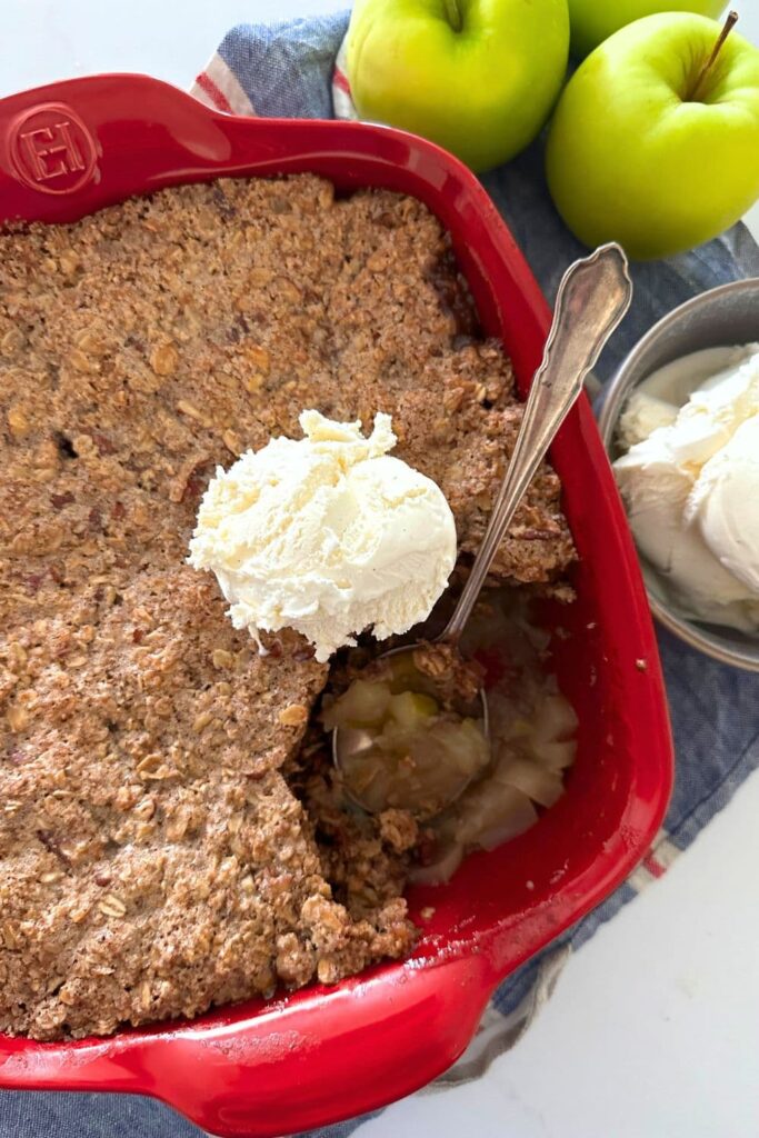 Sourdough apple crisp baked in square red baking dish. A piece of the apple crisp has been removed and placed into a bowl with vanilla ice cream and there are 3 green apples to the right of the baking dish.