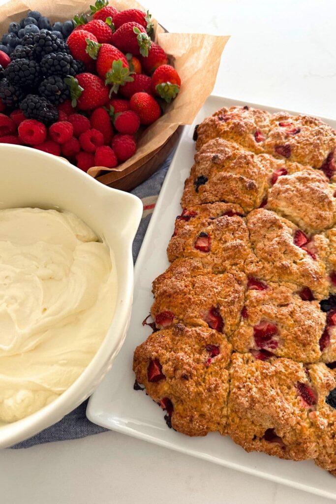A golden sourdough berry sheet pan shortcake sitting next to a bowl of vanilla whipped cream and bowl of summer berries.