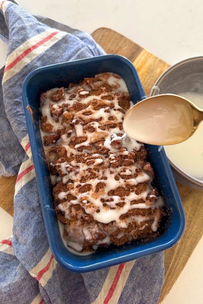 A loaf of sourdough discard apple fritter bread baked in a blue loaf pan. The sourdough apple fritter bread is being glazed with a vanilla glaze.