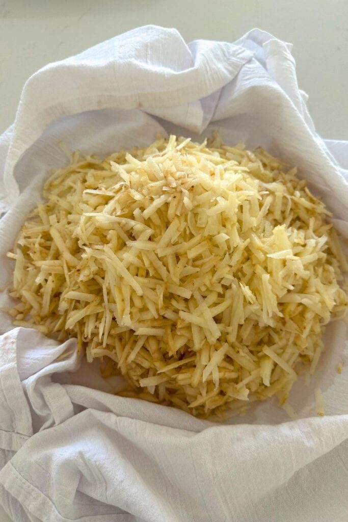 A pile of shredded potato sitting in a white flour cloth tea towel ready to make a sourdough hash brown casserole.