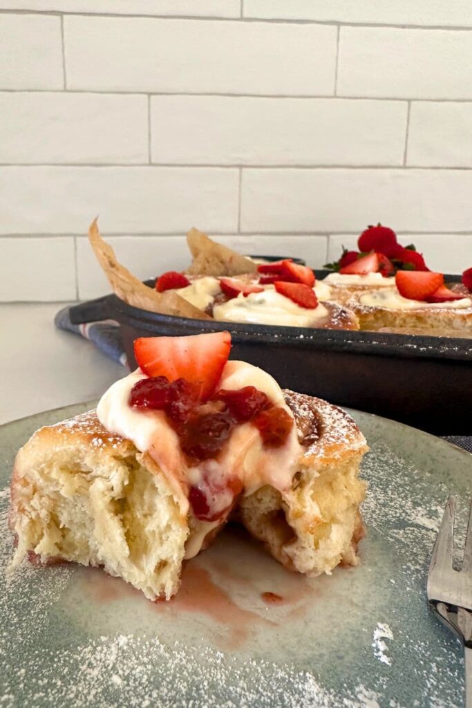 A sourdough strawberry roll sitting on a blue stoneware plate. There is a bite taken out of the sweet rolls so you can see the soft dough and bright red strawberry filling.