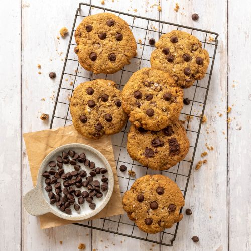 Six sourdough cookies with chocolate chips sitting on a wire cooling rack next to a small bowl of chocolate chips.