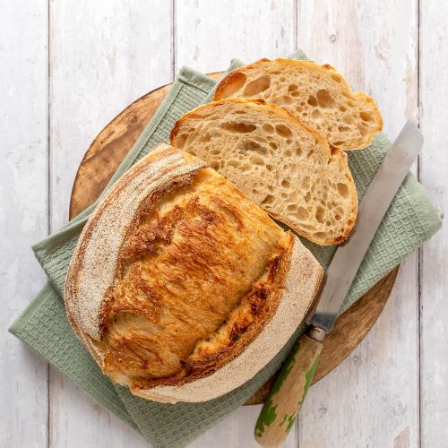 Sourdough loaf and 2 slices sitting next to a bread knife on a wooden cutting board covered with a sage green linen napkin.